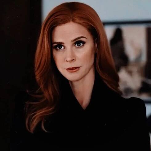 a woman with long red hair and blue eyes looks at the camera while wearing a black coat