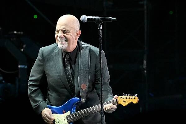 Billy Joel in a gray suit and playing a blue Fender Stratocaster guitar during a concert.