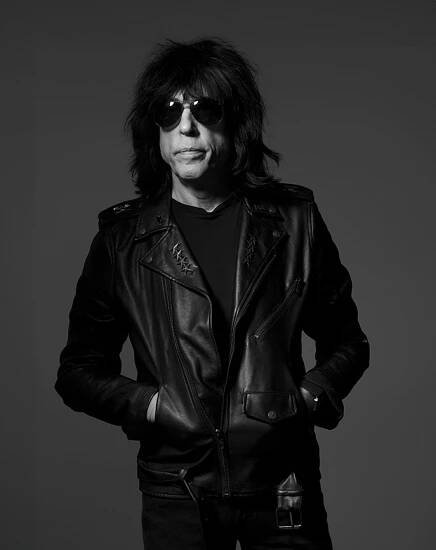 A black and white portrait of Marky Ramone by Albert Watson. He wears all black - a leather jacket, black pants and large sunglasses. His hands are in the pockets of his jacket. 