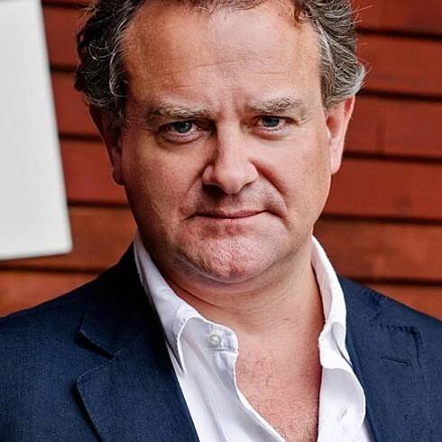 A close-up of Hugh Bonneville in a blue suit with white shirt, against a brown background