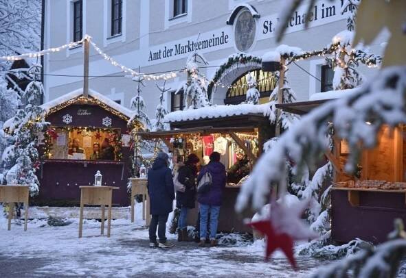 Verschneite Stände eines Weihnachtsmarktes im Salzburger Lungau, geschmückt mit Lichtern und Besuchern davor, am Abend.