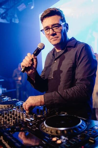 Andy Burnham wears glasses and a black shirt and looks into the camera holding a microphone while DJing on decks.