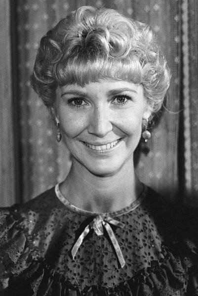 Person smiling, wearing a lace-detail, high-collared dress and earrings, with short, curled hair