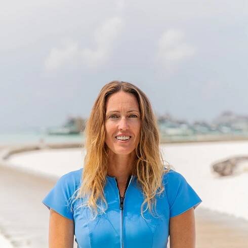 A woman with long wavy hair, wearing a blue short-sleeve wetsuit, stands and smiles on a sandy beach with a cloudy sky and water in the background.