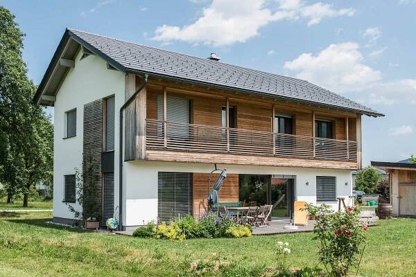 Modernes zweistöckiges WOLF Haus mit nachhaltiger Bauweise, Holzfassade und großer Terrasse im Garten, unter blauem Himmel.