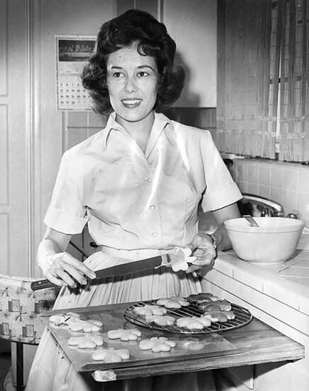 A black and white photo of a woman wearing a dress as she looks off to her right and spreads icing on a shamrock cookie with a knife, standing in a kitchen over a cutting board with shamrock cookies on a cookie sheet and cooling rack.