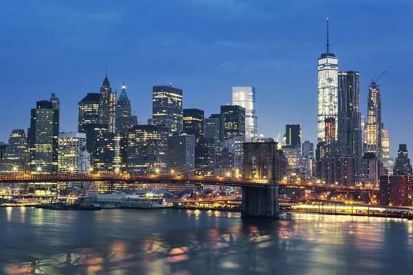 A city skyline at night with tall skyscrapers illuminated against a dark sky, reflecting on the water of a nearby lake or river. The scene captures the essence of a bustling metropolis with bright lights and urban architecture.