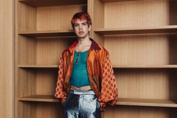Julio Torres, in shiny silver pants, green tank and orange shirt, stands with hands in pockets before empty shelves