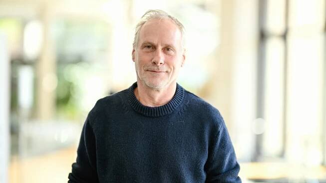 Der Schauspieler Wolfram Koch steht vor einer Pressekonferenz zum neuen Stück der Nibelungen-Festspiele. Er trägt einen dunkelblauen Pullover und schaut freundlich in die Kamera.