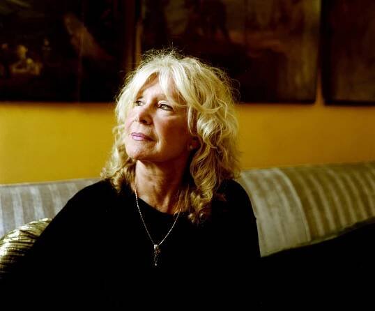 A portrait of Francine Pascal with blonde hair, a black top and a long necklace sitting on a sofa and looking wistfully away from the camera.