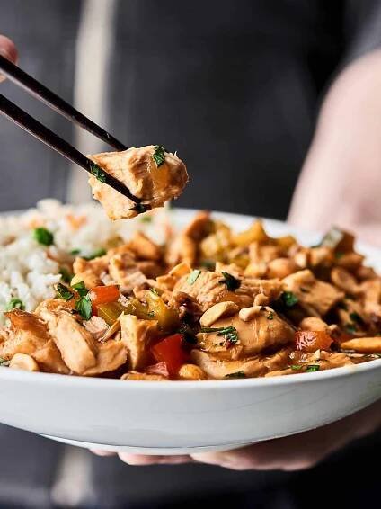 Piece of chicken being picked up with chopsticks plate of chicken and rice held