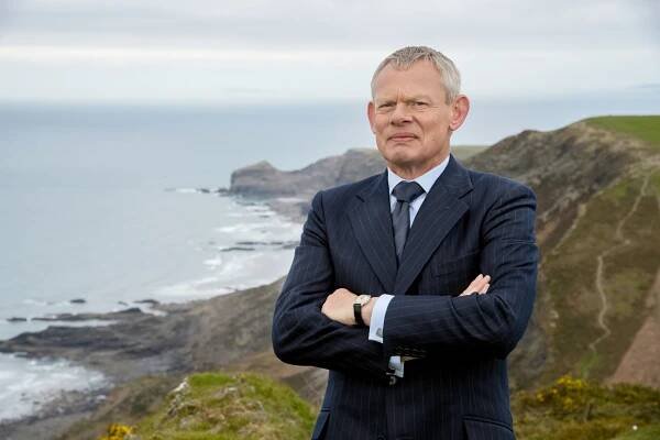 Martin Clunes as Doc Martin, arms crossed, standing on a cliff overlooking the ocean.