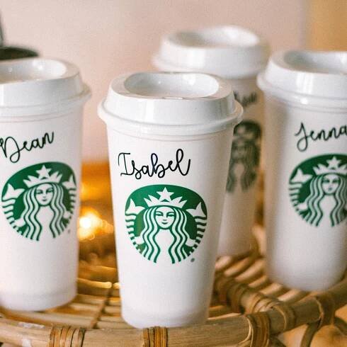 May include: Four white to-go coffee cups with green Starbucks logos and personalized names: Jean, Isabel, and Jenna. The cups are sitting on a woven wicker surface.