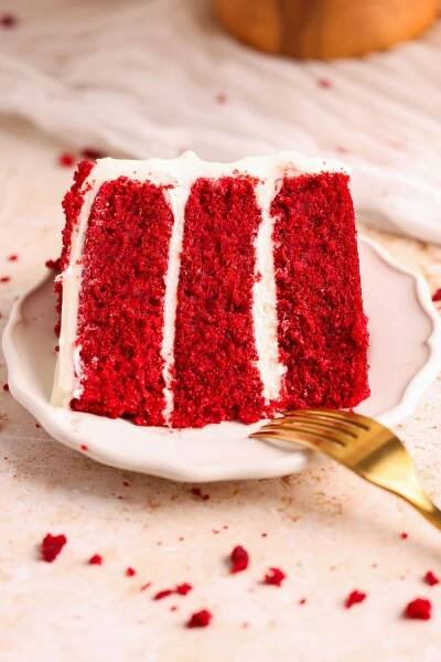 slice of red velvet cake on a small plate pictured from the side. the cake has three layers of moist soft cake with cream cheese frosting