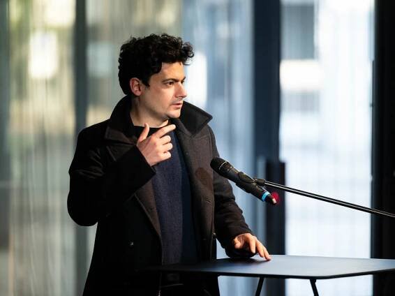 Clément Cogitore steht an einem Rednerpult und sprcht mit einer ausladenden Geste.
