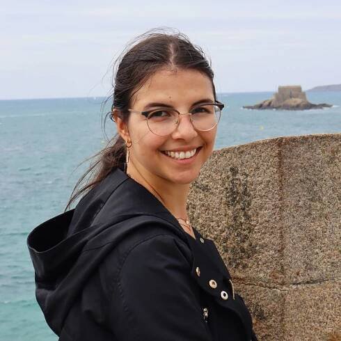 Junge Frau mit Brille, die an einer Mauer steht und am Wasser lächelt, im Hintergrund eine Felslandschaft.