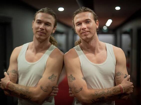 Young man wearing white singlet leans against mirror, in hallway.