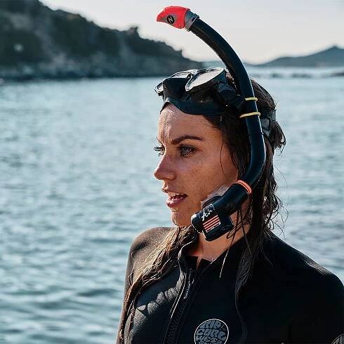 Frau im schwarzen Tauchanzug mit Taucherbrille und Schnorchel vor einer Wasserlandschaft.