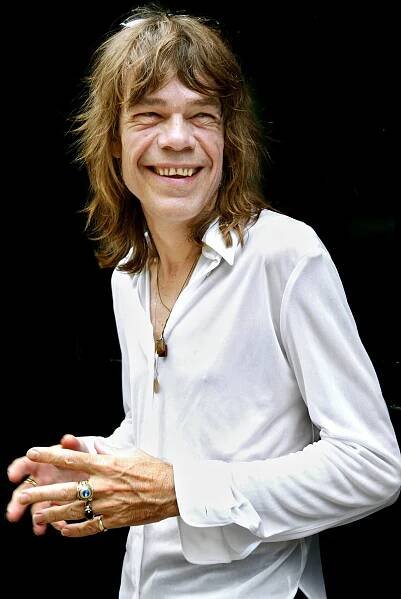 David Johansen, a skinny man with long brown hair wearing a long-sleeved white shirt, stands with is hands clasped in front of him and smiles broadly.