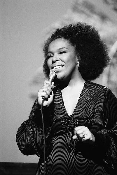A black-and-white photo of Roberta Flack, in a dark, patterned dress with a V-neck, singing into a microphone onstage.