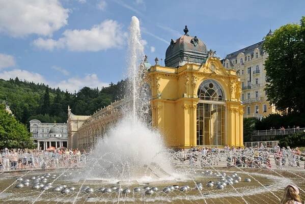 a fountain in front of a large yellow building with lots of people around it and water spouting from the top