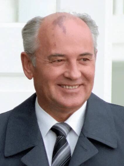 A man in a grey suit, white shirt and dark tie, balding with grey hair, he has a birthmark on his forehead