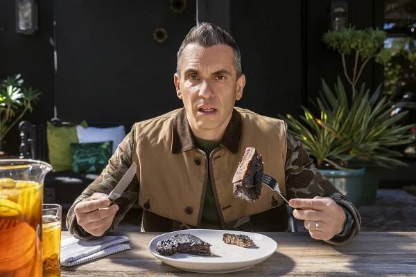 Photo of Sebastian Maniscalco sitting at a table and holding a hunk of steak with his fork.