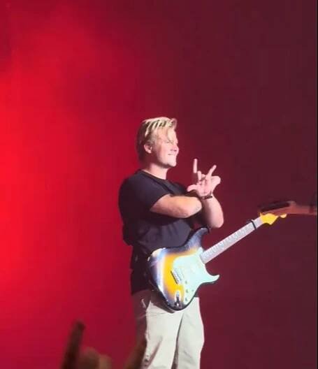 A musician on stage with a guitar, making a rock and roll hand gesture, under a red light with text 