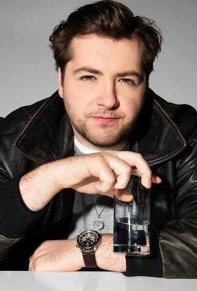 Michael Gandolfini wearing a brown leather jacket over a gray sweater holding a glass of water.
