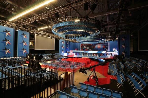 Audience view of the set for the Republican National Debates 2020 before the audience arrives. Red carpet with blue chairs in front of a mostly blue stage that has red, white, and blue colored stars on a blue backdrop. A video screen is behind the lecterns where the candidates will stand. Two sets of circular trusses on diminishing side hang one inside the other over the audience. Lights and a circular banner with the same multi-colored stars hang from the circle trusses. TV cameras are set up in different areas of the venue.