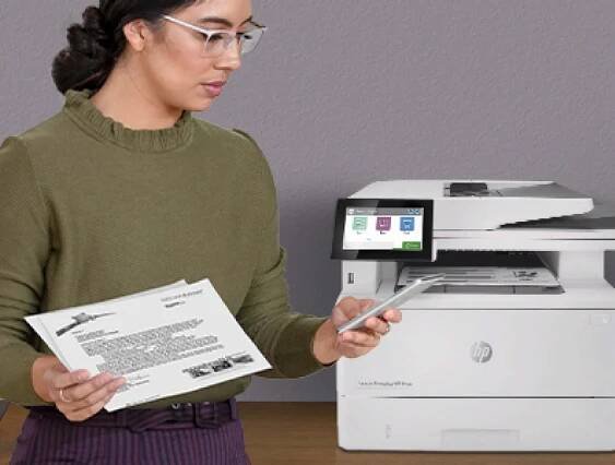 Žena u multifunkční tiskárny HP Color LaserJet Enterprise drží vytištěné dokumenty a používá aplikaci Workflow na mobilním telefonu