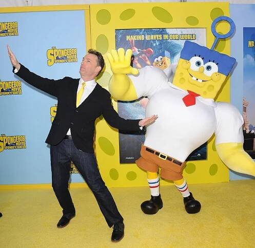 Tom Kenny playfully poses next to a life-sized SpongeBob character in front of a SpongeBob movie promo backdrop