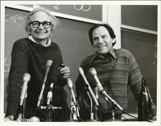 Black and white photo of Torsten Wiesel (left) and David Hubel (right) smiling and standing at a podium with microphones and a bottle of champagne.