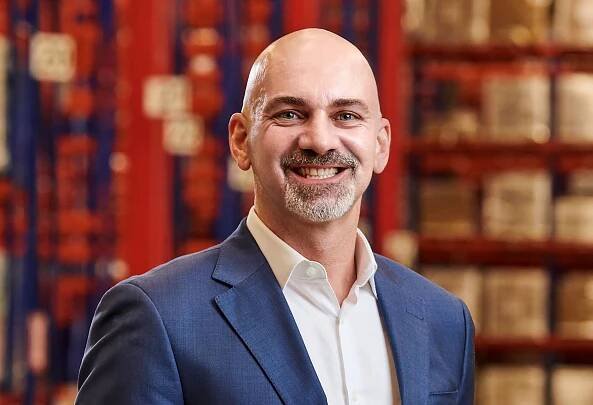 A man with a gray goatee, wearing a blue suit and white shirt, smiles at the camera. He stands in front of shelves with various boxes and items, suggesting a warehouse or storage facility background.
