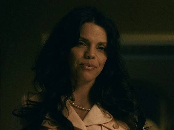 vanessa ferlito as carmen in griselda, wearing a pink shirt and with her hair worn curled and loose. she's pursing her lips