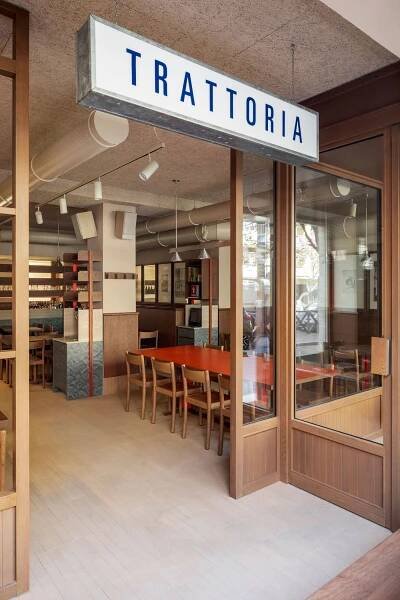 The entrance to Santo Palato in Rome, designed by Naessi Studio, under a prominent "TRATTORIA" sign. Large wooden-framed glass doors open into a bright, inviting space. Inside, a vibrant orange communal table is visible, surrounded by minimalist wooden chairs, hinting at the modern yet authentic dining experience within.