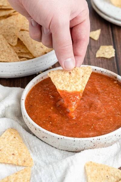 a hand dipping a tortilla chip in a bowl of fresh salsa roja.