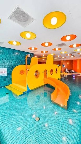 Crystal Mountain Hotel Wisła Zewnętrze zdjęcie The photo shows a brightly colored children's water play area in a swimming pool. The main feature is a large, yellow and orange play structure shaped like a boat or submarine. It has several circular openings and a curved orange slide extending from