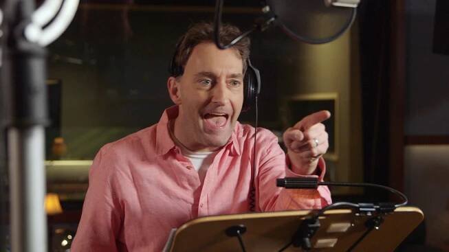 Person in a recording studio, wearing headphones and a casual shirt, gestures energetically toward a microphone
