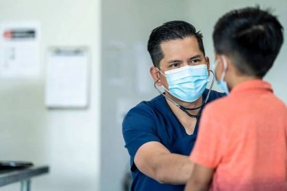 Mužský lékař v masce a rukavicích při kontrole u mladého, 8letého chlapce kvůli vypuknutí COVID-19.