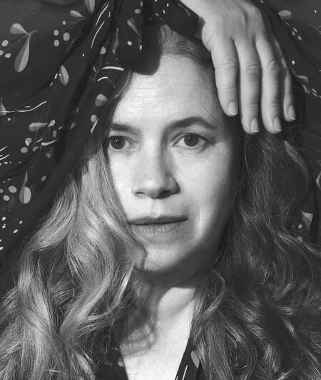 A beautiful portrait of Natailie Merchant. She is staring directly out and her face is framed by her hand over her head and her long hair on either side. The hair cascades over a part of her face on her right side.