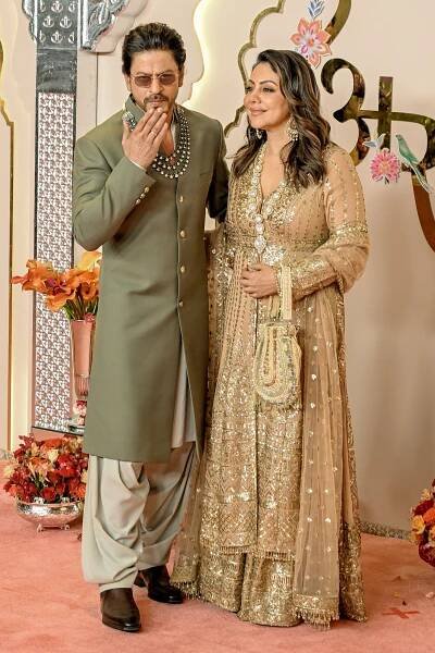 Shah Rukh Khan and his wife, Gauri Khan, posing for paparazzi on a red carpet. They are wearing olive-green and gold formal Indian attire, and he is blowing a kiss toward the cameras.