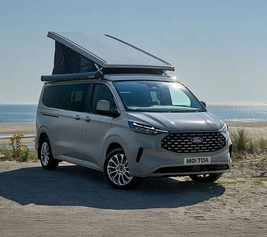 Stříbrný Ford Transit Custom Nugget s otevřenou střechou zaparkovaný na pláži u moře.