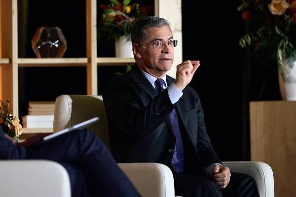 Secretary of Health and Human Services Xavier Becerra speaks at a media conference. He is seated in a cream arm chair and speaks to the audience. He wears a dark blue suit, tie and thin-rimmed glasses.