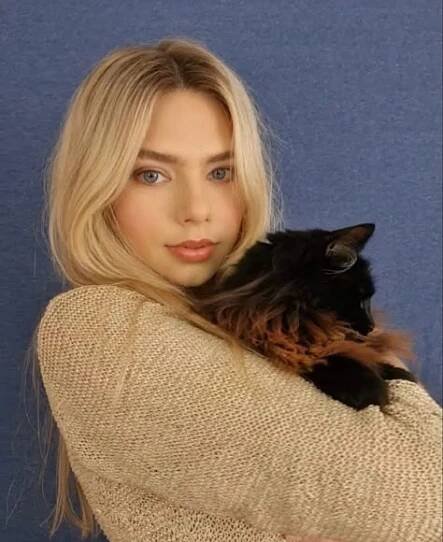 a woman holding a cat in her arms with a blue wall behind her and she is wearing a tan sweater