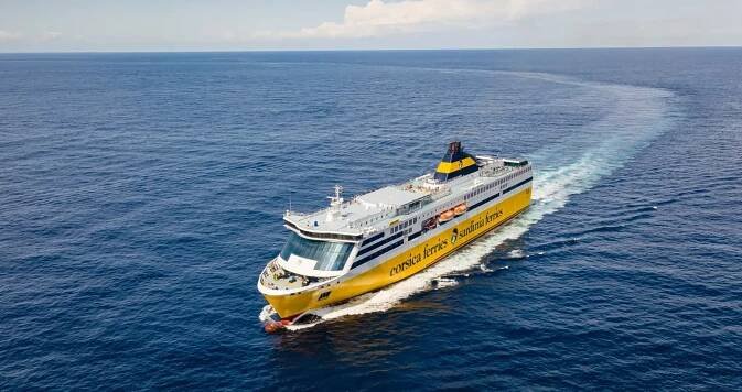 The Mega Express passenger ferry by Corsica Ferries sails across the sea under a clear sky. The vessel is painted yellow with a white superstructure and moves toward the right side of the image.