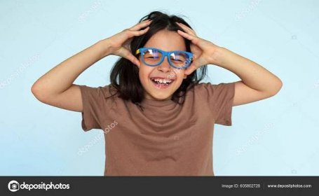 Stáhnout - Studio portrait of a pretty joyful little girl smiling broadly wearing a brown t-shirt, blue eyeglasses  enjoying a good mood holds her hands on her head isolated on blue background. — Stock obrázek