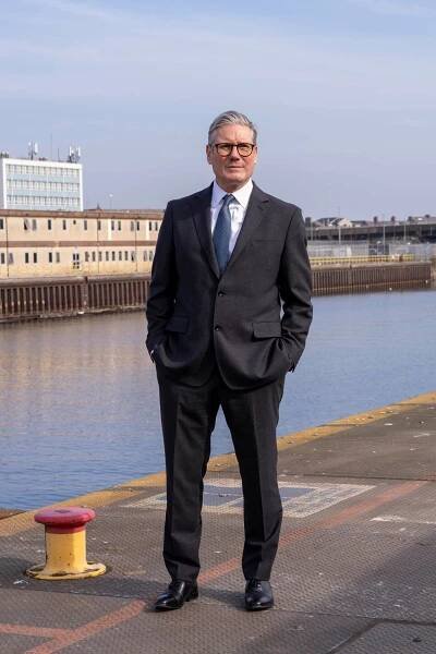A man wearing a suit stands with his hands in his pockets on a dock.