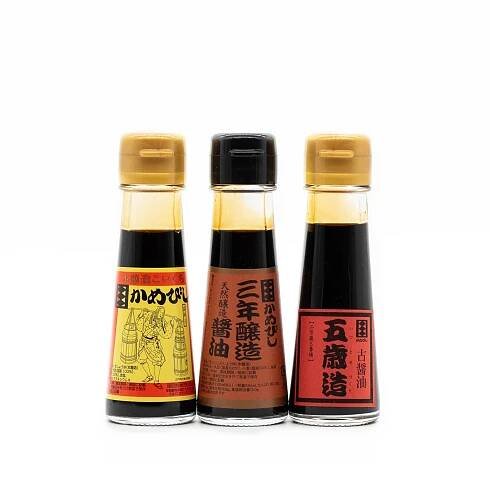 The Aged Soy Sauce Triple Pack features three premium glass bottles with gold caps and red and black labels, displayed side by side against a white background, highlighting their renowned rich umami flavor.