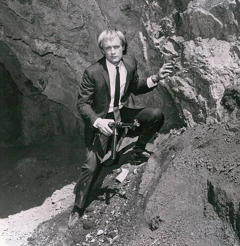 A black-and-white photo of David McCallum, a young man in a suit and tie with blond hair in bangs, standing on a rock formation with his left hand on a rock and a gun in his right hand.